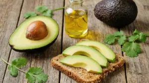 Avocado a metà e fette su pane integrale con una bottiglietta di olio e foglie di coriandolo su tavolo in legno
