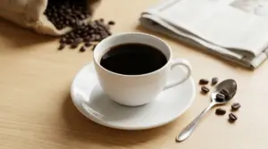 Tazza di caffè nero amaro su piattino bianco con chicchi di caffè e cucchiaino su tavolo di legno