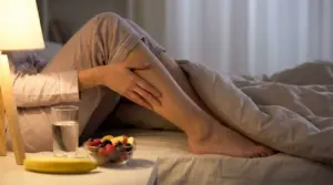 Persona seduta sul letto di notte si massaggia il polpaccio per un crampo alle gambe, con acqua e frutta sul comodino