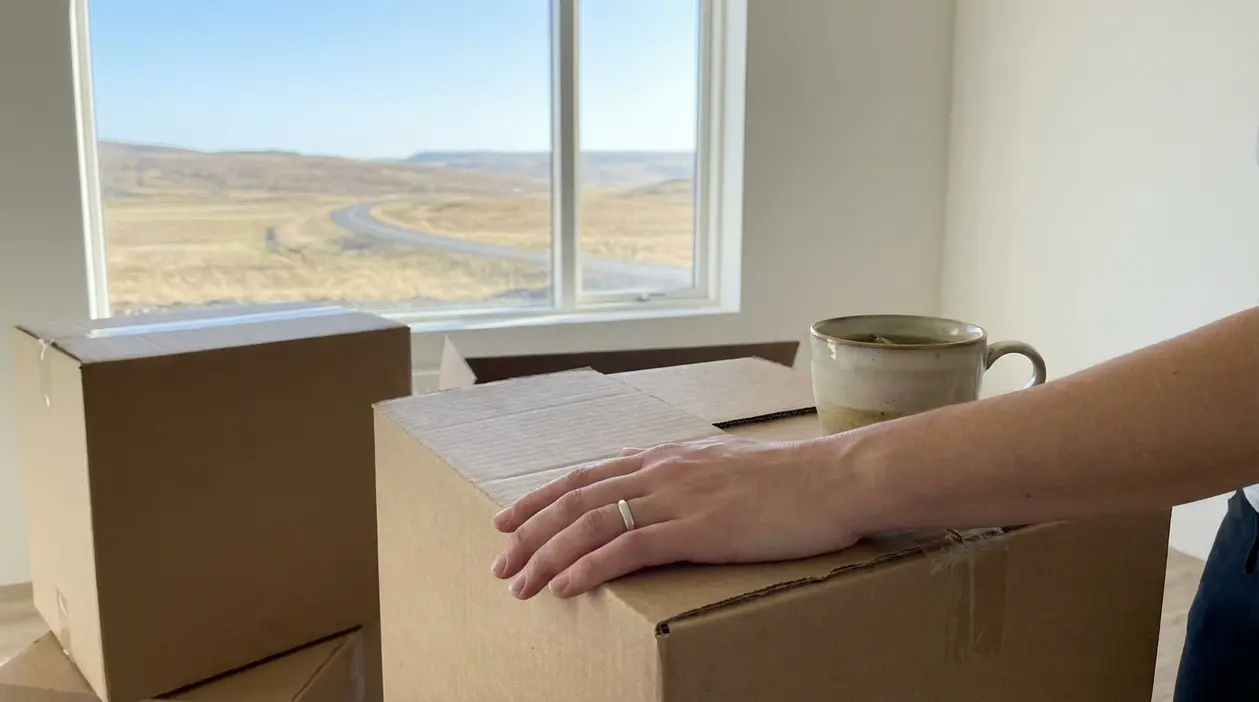 Una persona con la mano su scatoloni da trasloco in una stanza vuota, davanti a una finestra