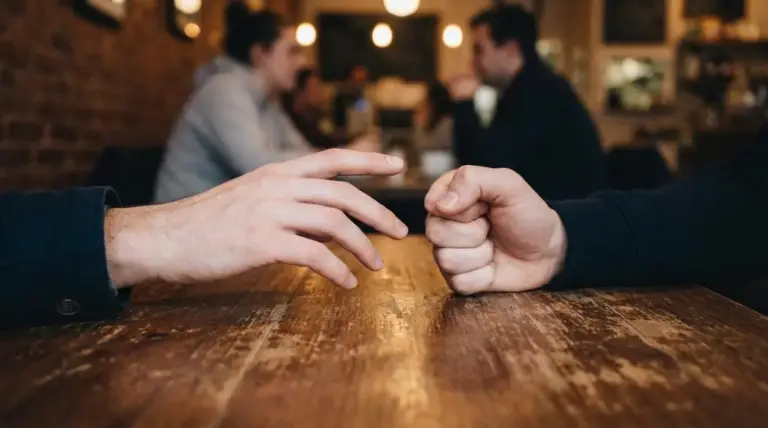 Due persone al tavolo in un bar discutono: una mano indica e l'altra è chiusa a pugno, segno di tensione