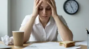 Persona seduta alla scrivania con le mani sulla testa, stanca e stressata, con documenti e orologio sullo sfondo