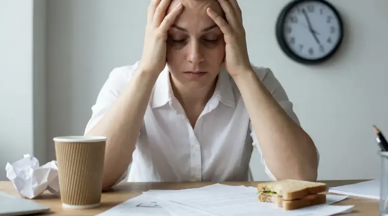 Persona seduta alla scrivania con le mani sulla testa, stanca e stressata, con documenti e orologio sullo sfondo