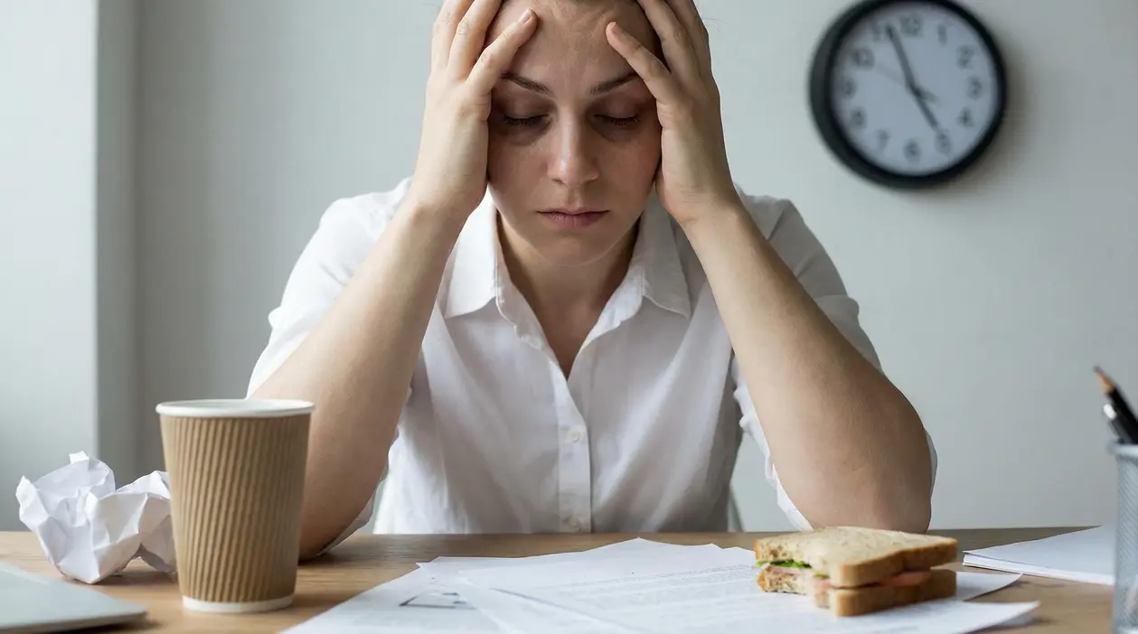 Persona seduta alla scrivania con le mani sulla testa, stanca e stressata, con documenti e orologio sullo sfondo